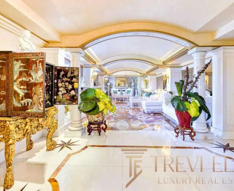 Роскошный интерьер роскошного дома с классическими элементами, белыми колоннами, золотой мебелью и зелеными растениями, создающими элегантную атмосферу.