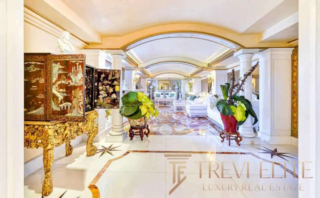 Роскошный интерьер роскошного дома с классическими элементами, белыми колоннами, золотой мебелью и зелеными растениями, создающими элегантную атмосферу.