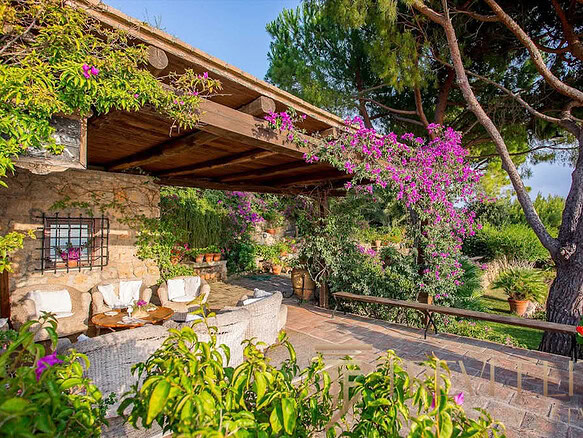 Уединенная терраса в итальянском стиле с цветущими растениями и уютной зоной отдыха под деревянным навесом, окруженная зеленью и пышной растительностью.