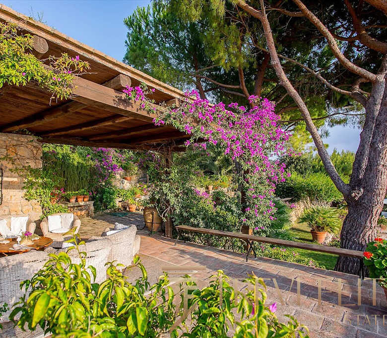 Уединенная терраса в итальянском стиле с цветущими растениями и уютной зоной отдыха под деревянным навесом, окруженная зеленью и пышной растительностью.