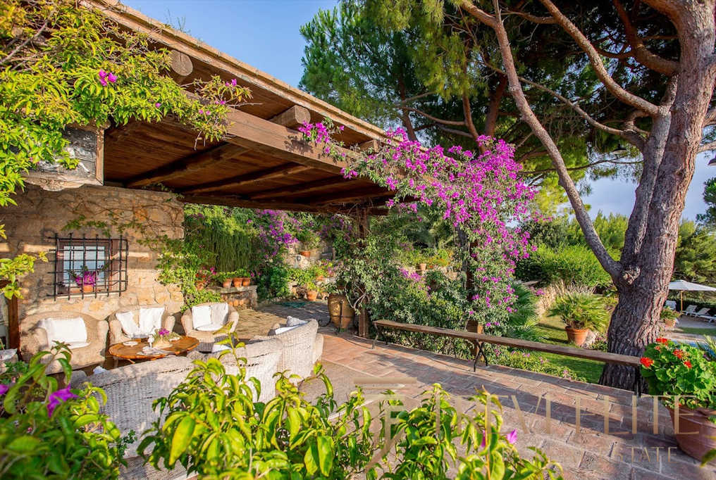 Уединенная терраса в итальянском стиле с цветущими растениями и уютной зоной отдыха под деревянным навесом, окруженная зеленью и пышной растительностью.