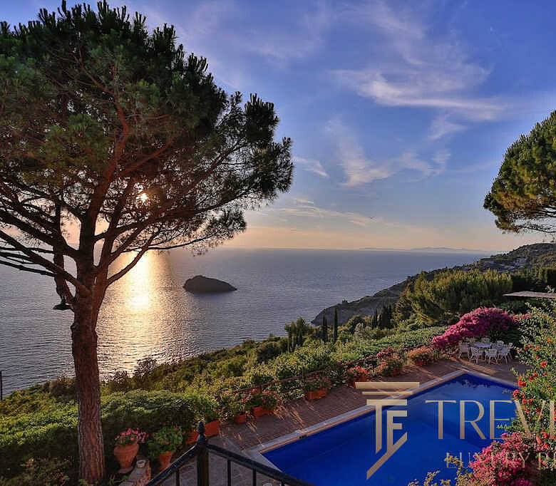 Узкий вид на бассейн и зеленый сад на фоне морского побережья во время заката, окруженный высокими деревьями и цветущими кустами, демонстрирующий роскошные особенности недвижимости Τреви Элит.