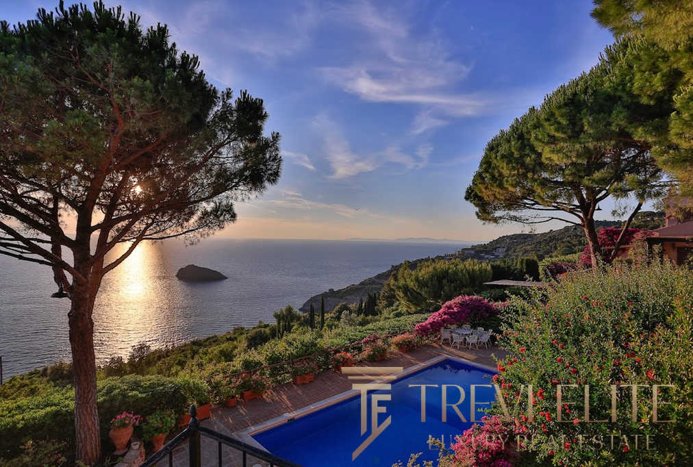 Узкий вид на бассейн и зеленый сад на фоне морского побережья во время заката, окруженный высокими деревьями и цветущими кустами, демонстрирующий роскошные особенности недвижимости Τреви Элит.