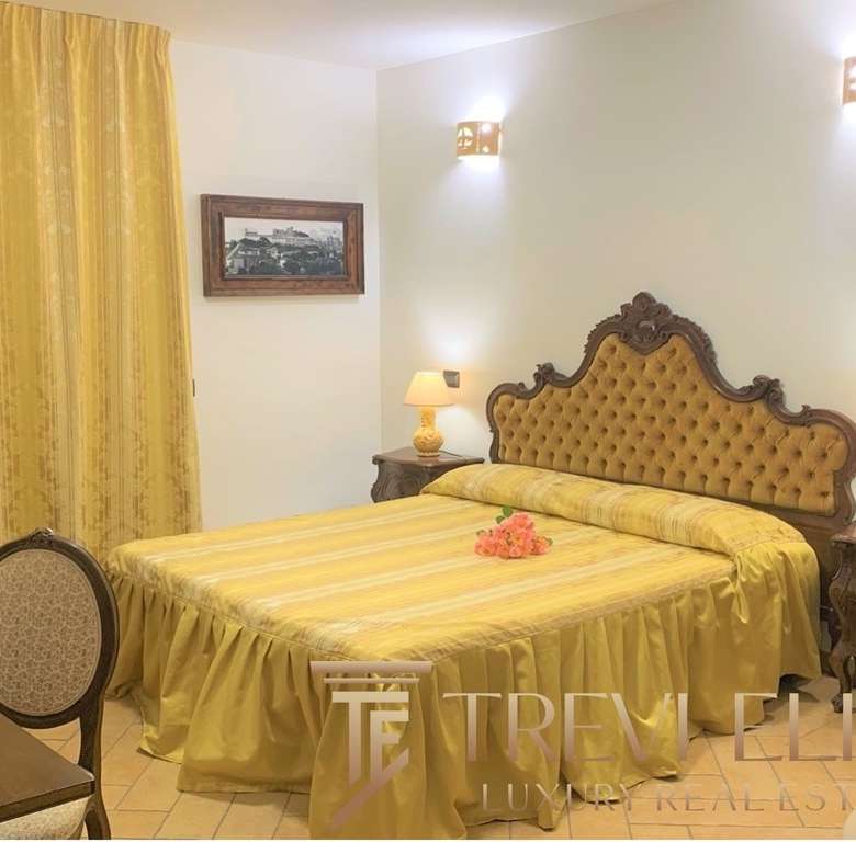 Элегантная спальня в классическом стиле с желтым постельным бельем, мягкой обивкой на изголовье кровати и деревянной мебелью, уютное освещение и картина на стене создают роскошную атмосферу.