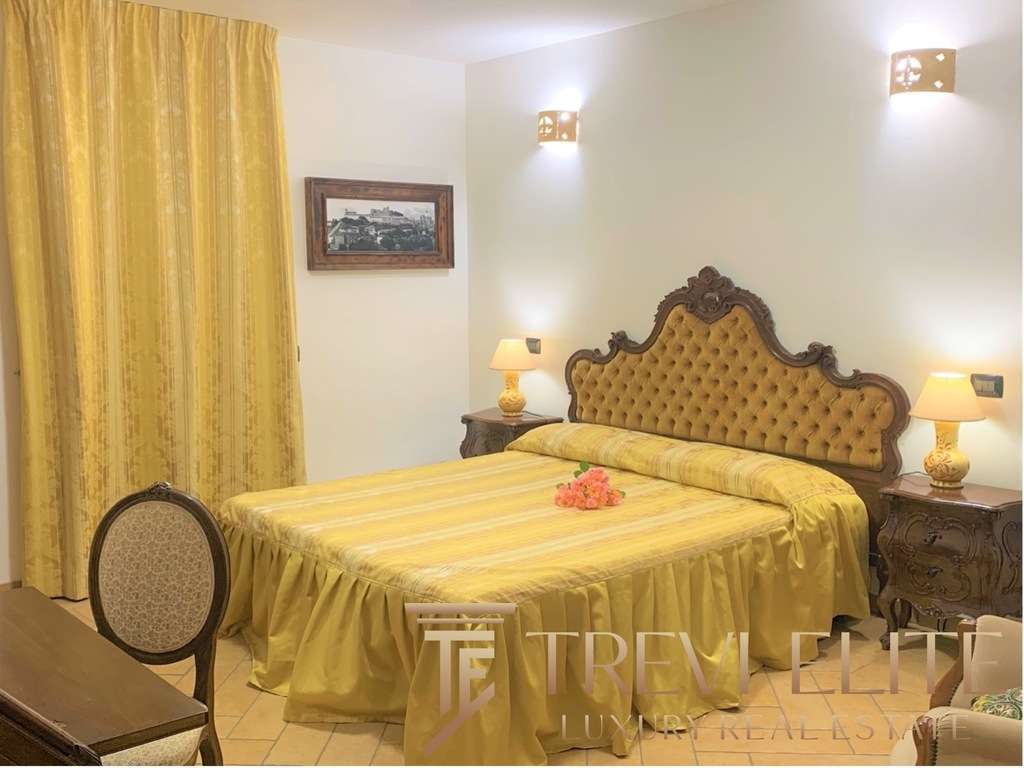 Элегантная спальня в классическом стиле с желтым постельным бельем, мягкой обивкой на изголовье кровати и деревянной мебелью, уютное освещение и картина на стене создают роскошную атмосферу.