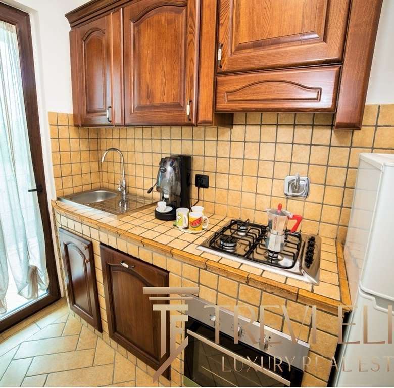 Угловая кухня с деревянными шкафчиками и плитой, рядом холодильник и окна с лёгкой шторой. Просторное и уютное пространство для кухни с современными аксессуарами и классическим стилем.