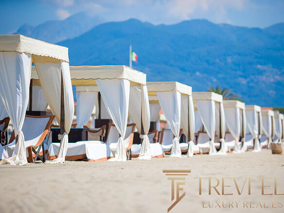 Роскошные пляжные шезлонги с белыми навесами на песчаном берегу моря на фоне гор, создающие атмосферу элитного отдыха и эксклюзивных курортных услуг.