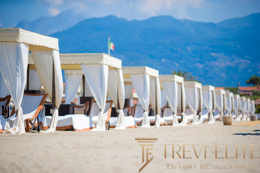 Роскошные пляжные шезлонги с белыми навесами на песчаном берегу моря на фоне гор, создающие атмосферу элитного отдыха и эксклюзивных курортных услуг.