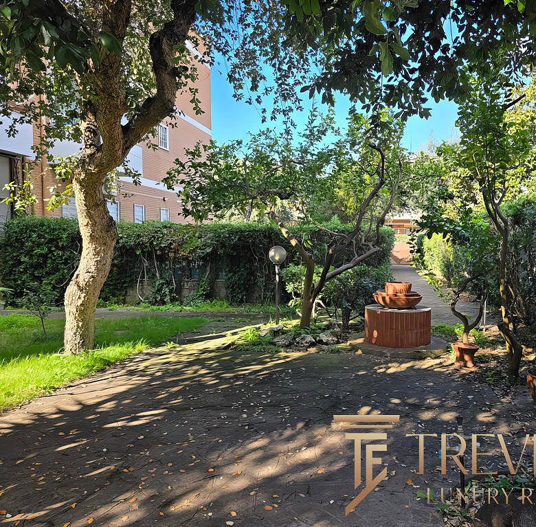 Уютный зеленый двор с деревьями и садовой мебелью у жилого комплекса Треви Элит. В тени создается спокойная атмосфера для отдыха или прогулок на свежем воздухе.