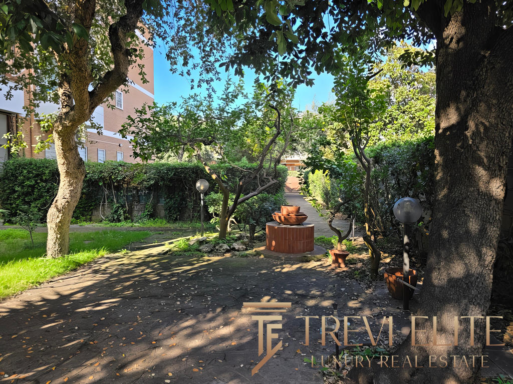 Уютный зеленый двор с деревьями и садовой мебелью у жилого комплекса Треви Элит. В тени создается спокойная атмосфера для отдыха или прогулок на свежем воздухе.