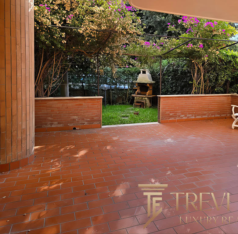 Уютная терраса с кирпичным полом и видом на зеленый сад с цветущими кустами и уличной печью для барбекю, идеальное место для отдыха на природе.