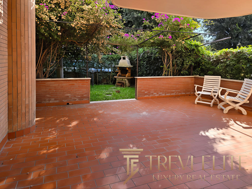 Уютная терраса с кирпичным полом и видом на зеленый сад с цветущими кустами и уличной печью для барбекю, идеальное место для отдыха на природе.