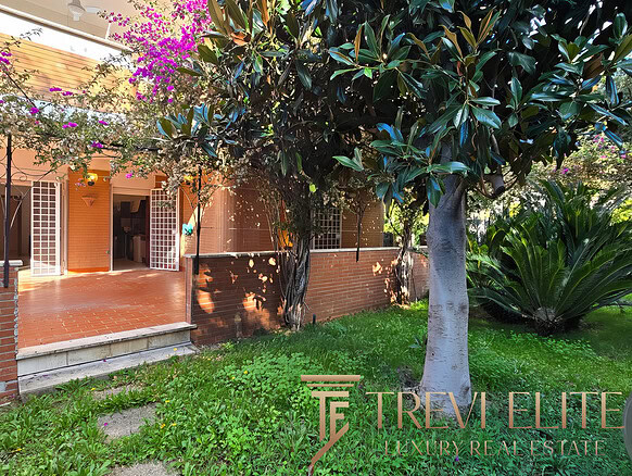 Изображение зеленого сада с деревьями и уютной террасой на заднем дворе дома. На заднем плане видны открытые двери, ведущие в жилое помещение, окруженное пышной растительностью и тенистыми деревьями.