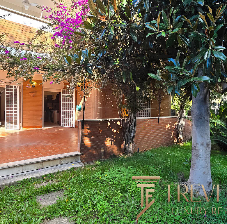 Изображение зеленого сада с деревьями и уютной террасой на заднем дворе дома. На заднем плане видны открытые двери, ведущие в жилое помещение, окруженное пышной растительностью и тенистыми деревьями.