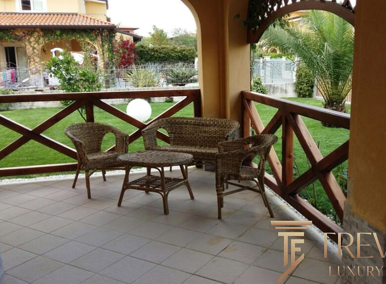 Уютное деревянное кресло и стол на террасе с видом на зеленый сад и соседние коттеджи. Идеальное место для отдыха и наслаждения природой. Надежное ограждение из дерева защищает пространство.