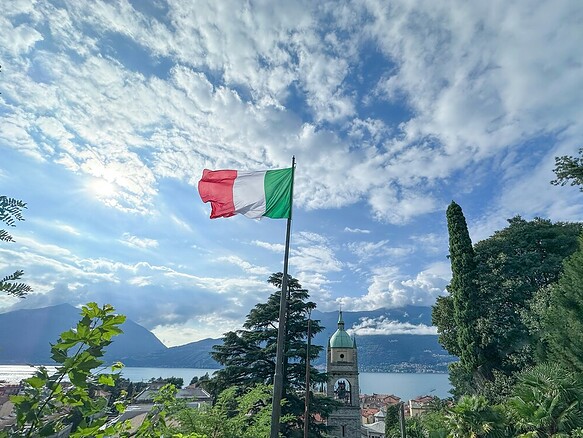 Итальянский флаг на фоне оживленного городского пейзажа с озером и горами.